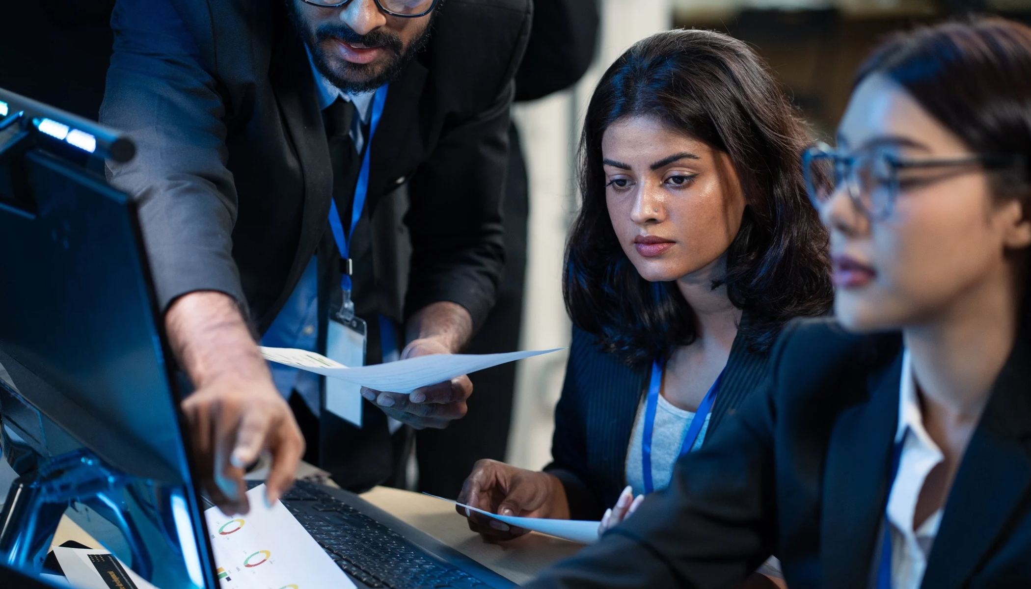 Three people in business attire look at a computer screen: a person with glasses and a beard (left), a person with shoulder-length hair (center), and a person wearing glasses with their hair in a bun (right).